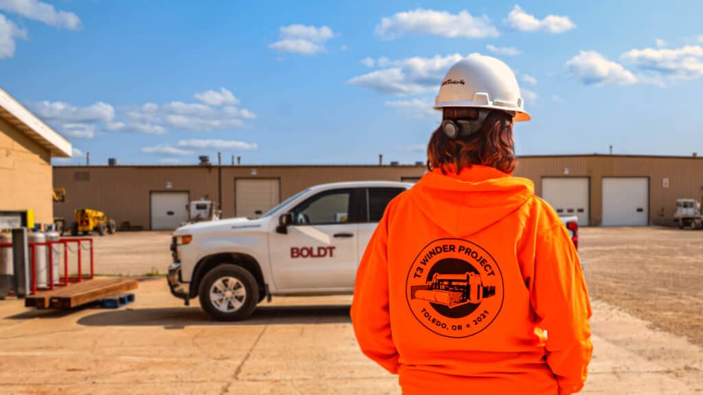 Construction worker wearing blaze orange and hard hat standing by construction truck outside project site