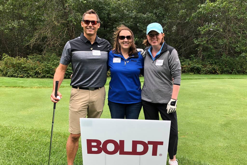 3 participants in the Golf for Research event - standing at a Boldt sign