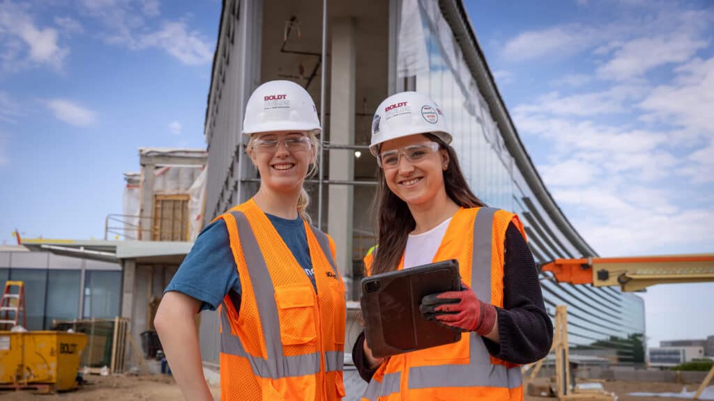 Two young construction women in safety vests and hard hats standing in front of large construction site building